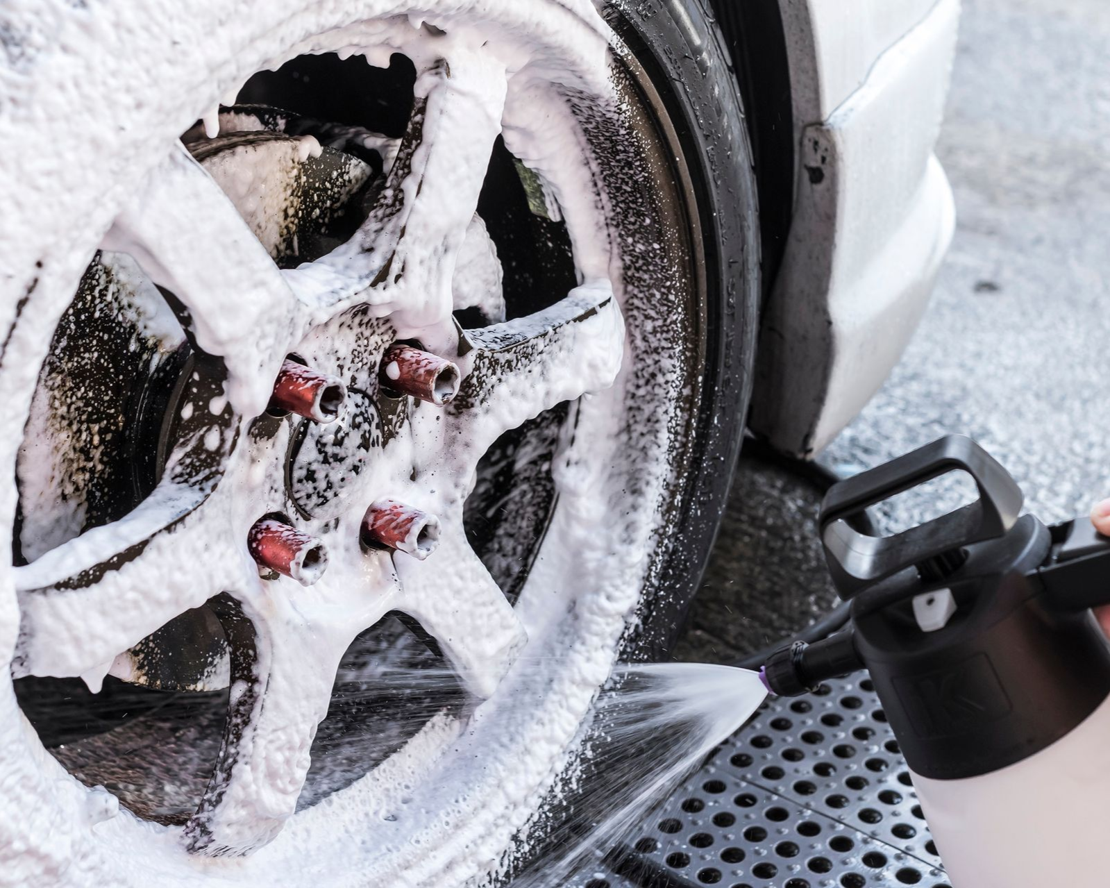 Foamy white soap being sprayed onto a car's alloy wheel during a wash.