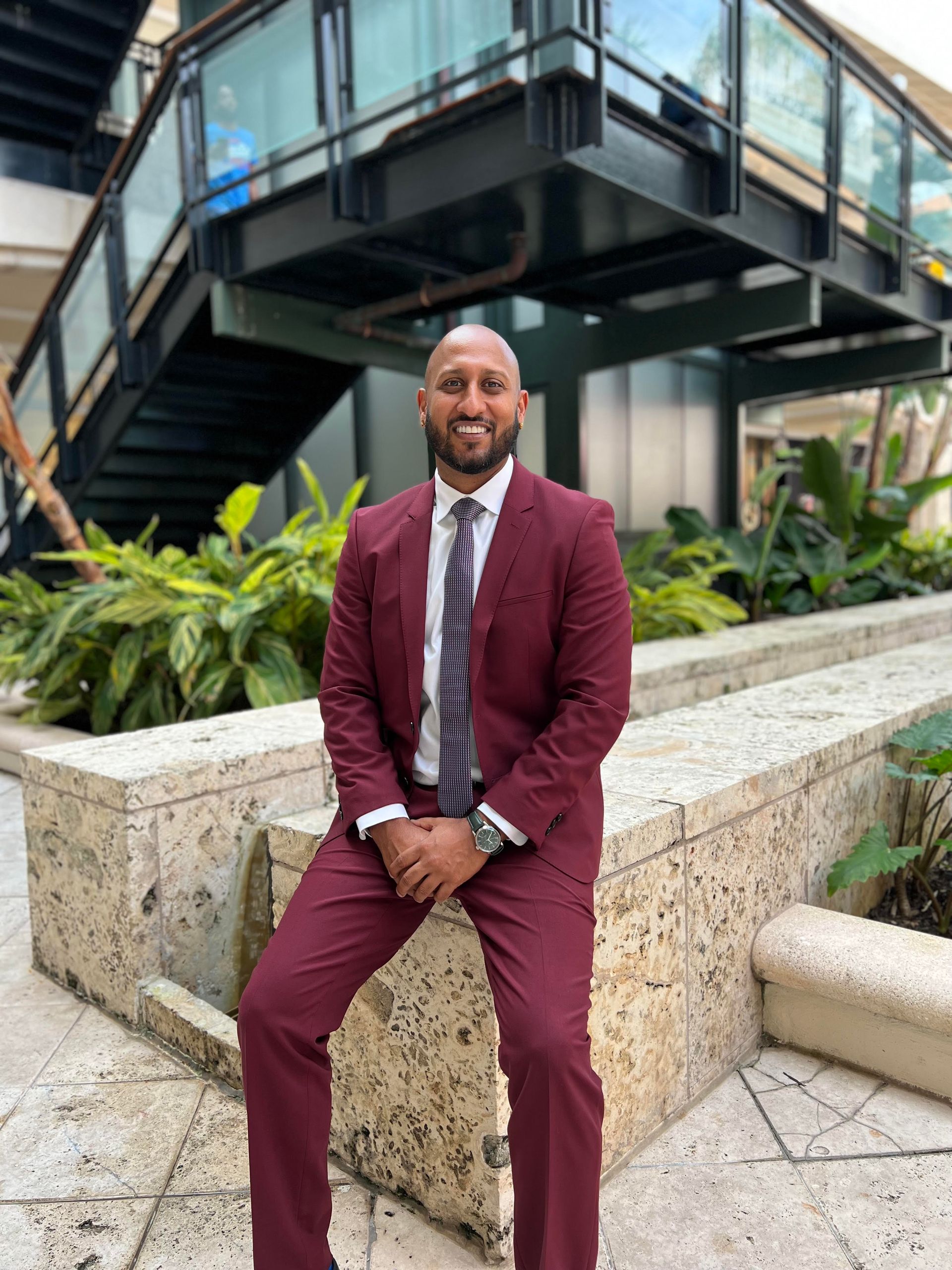 Man in burgundy suit sits on a stone wall outside, smiling.