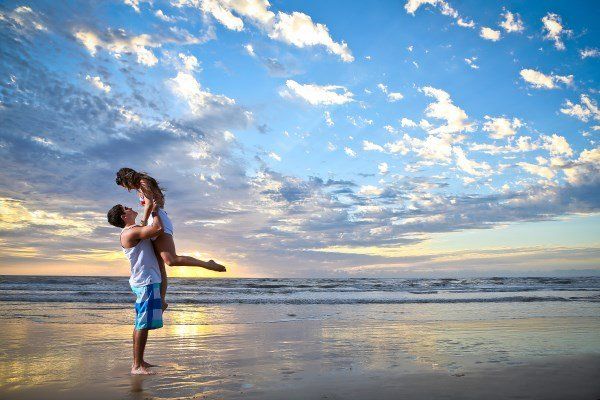 Um homem está segurando uma mulher nos braços na praia ao pôr do sol.