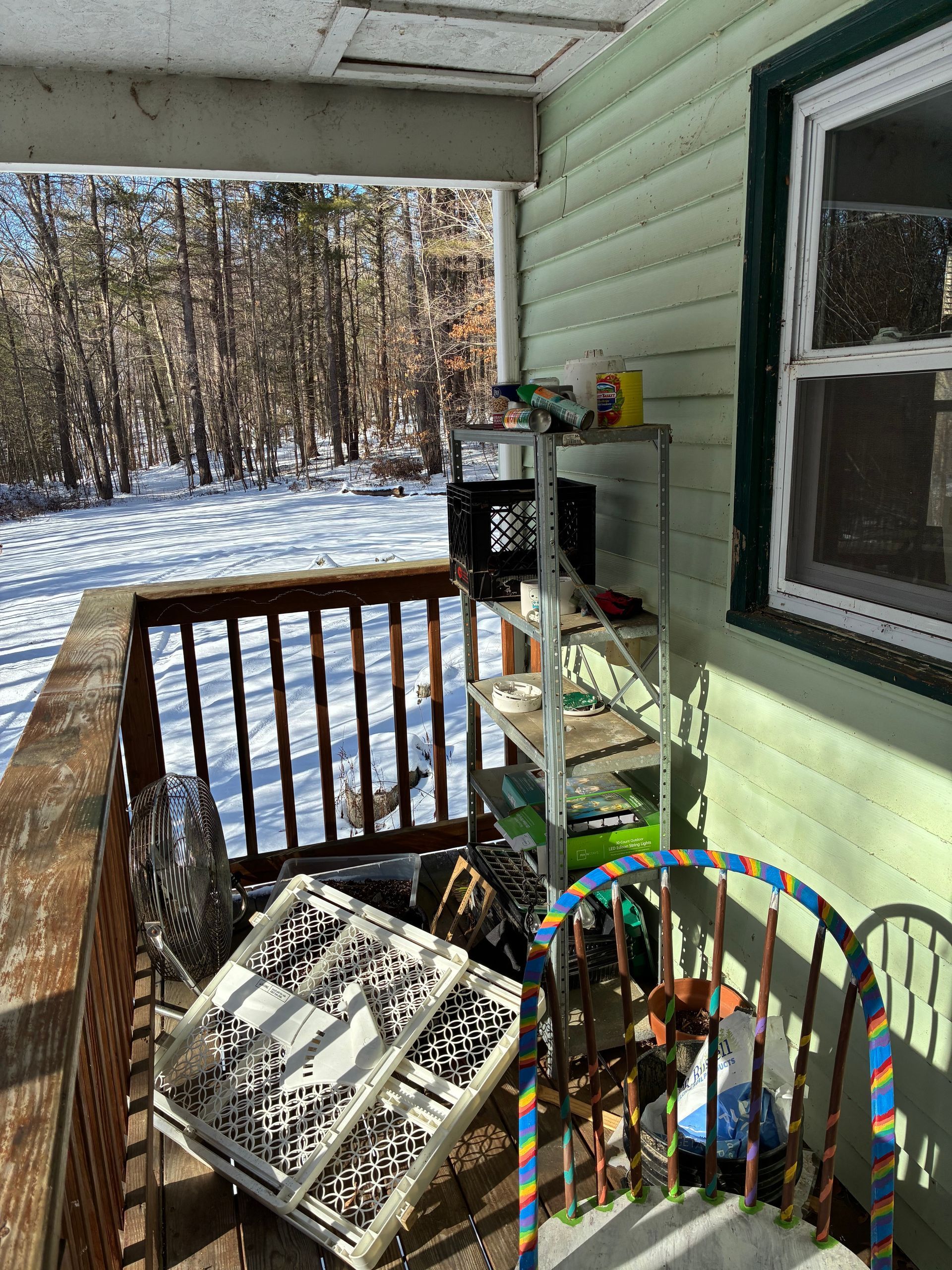 Porch with snow, a shelf unit, and miscellaneous items; a wooden railing and a window are visible.