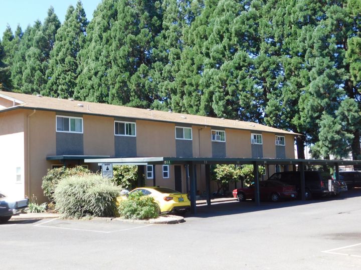 Apartment building with covered parking; tan walls, cars parked below, tall green trees in the background.