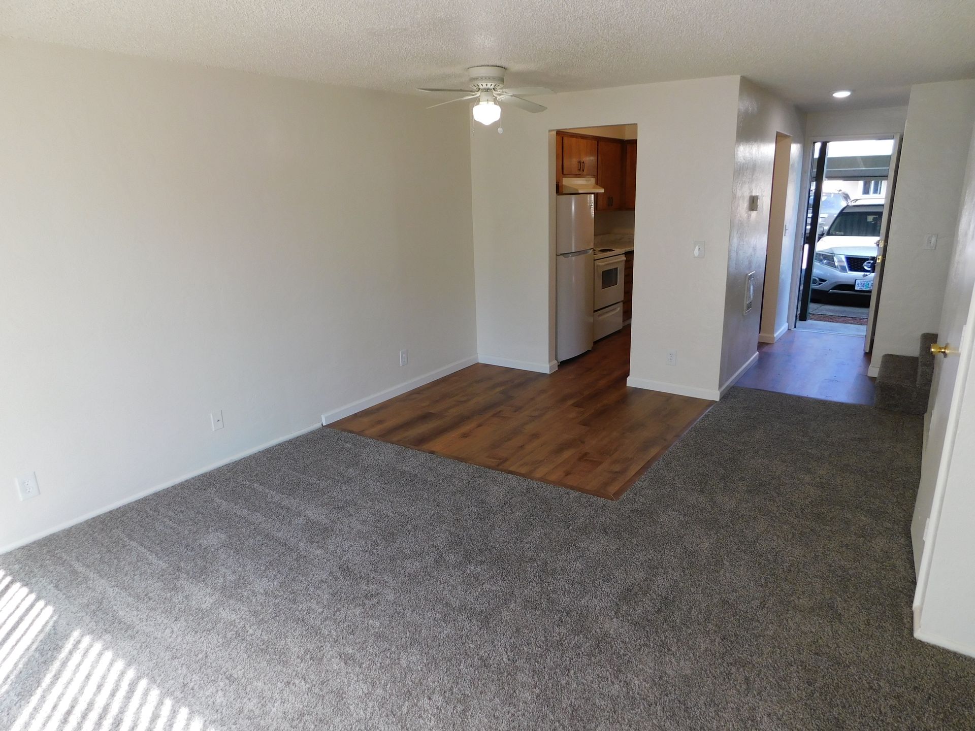 An apartment interior with gray carpet, wood flooring, and a view into the kitchen and hallway.