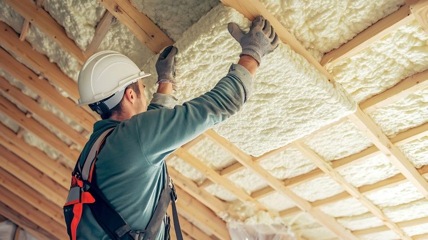 Construction worker installing insulation in an attic. Beige foam insulation, wooden beams, white hard hat.