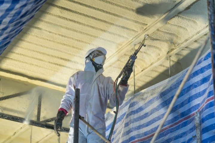 Person in protective suit spraying insulation on a ceiling, outdoors.