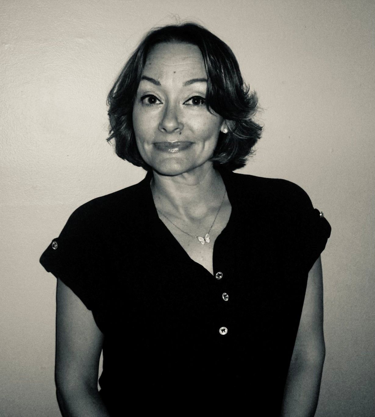 Woman with short dark hair in a black top, smiling slightly, against a light background.