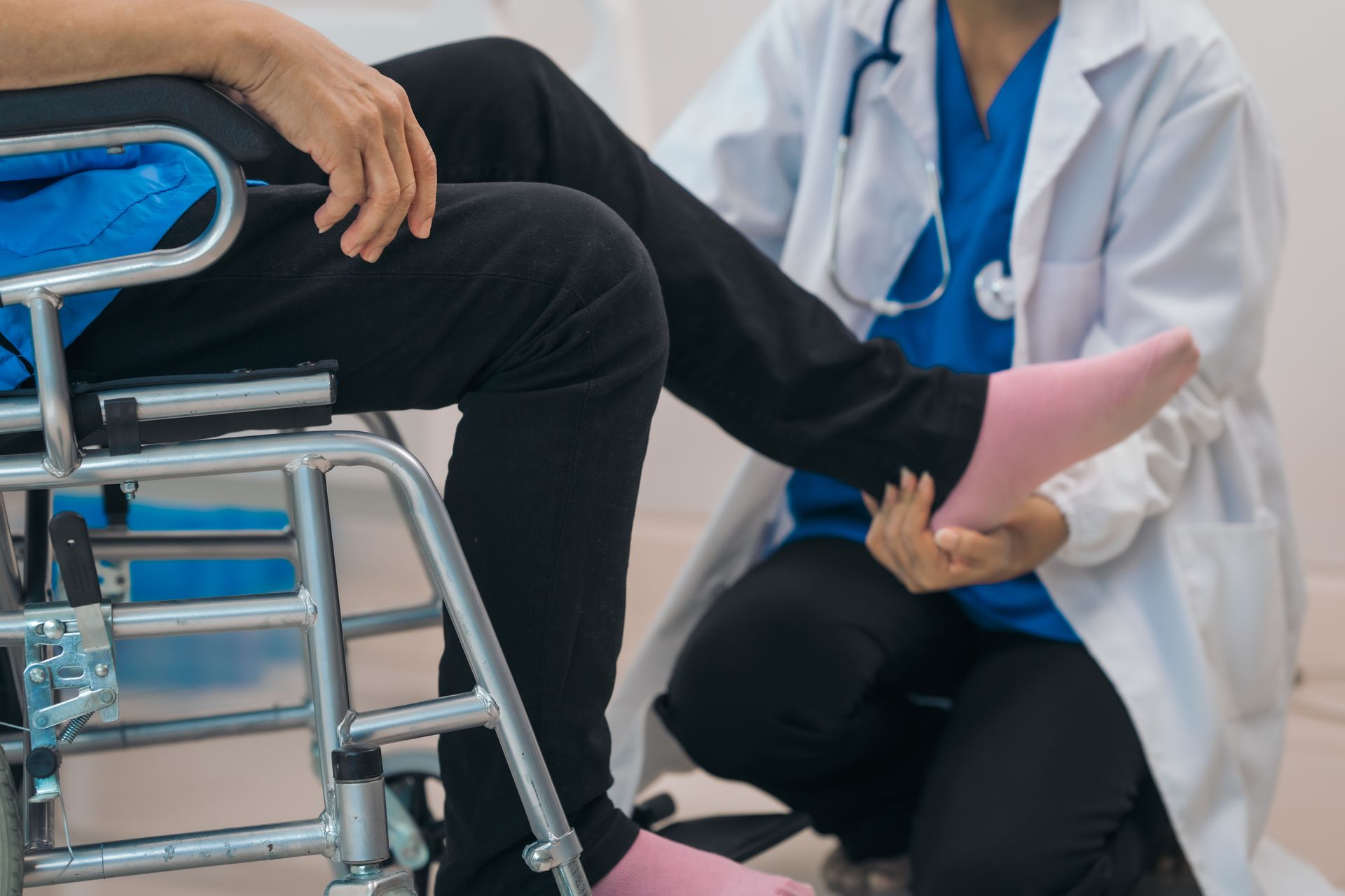 Doctor examining patient's leg in a wheelchair. The doctor wears a lab coat, patient has a blue shirt and pink sock.