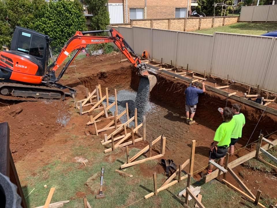 An eXcavator Pours Concrete Into a Trench, With Workers, Forms, and a Fence in a Backyard — Banora Pools in Tweed Heads South, NSW