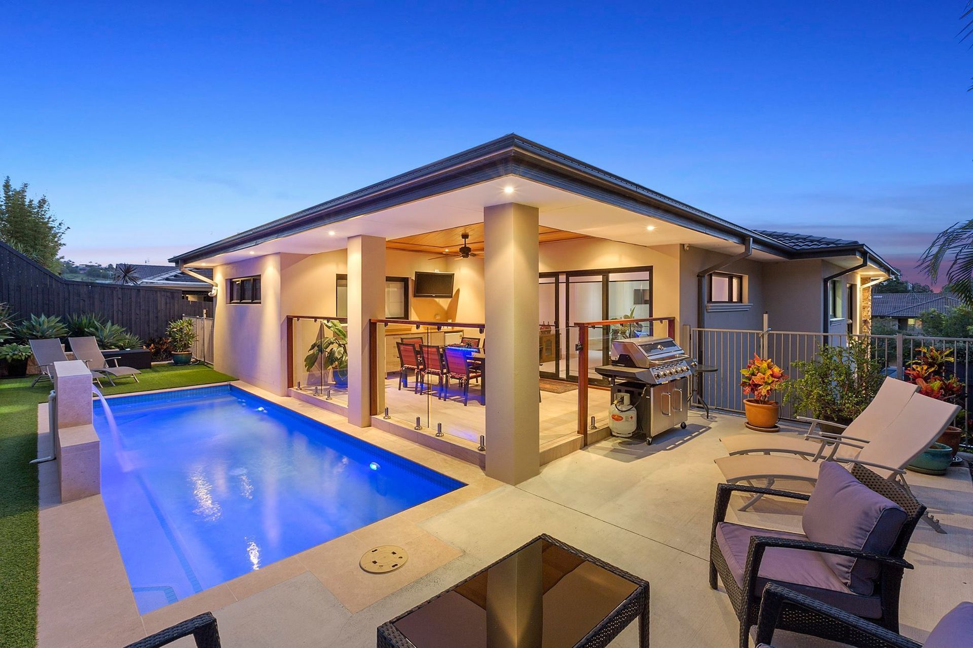 Backyard with pool, outdoor kitchen, and seating under a modern home with blue sky at dusk. — Banora Pools in Tweed Heads South, NSW