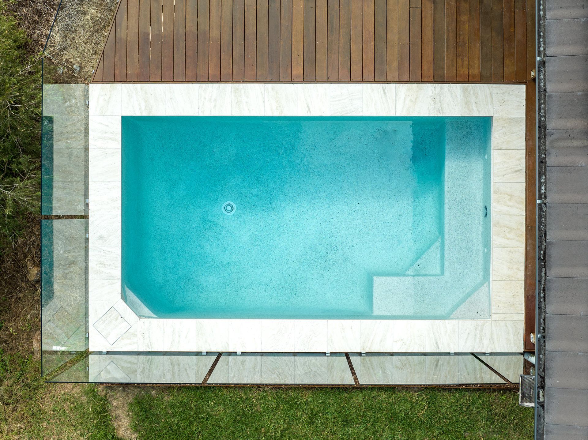 Aerial view of a rectangular swimming pool with turquoise water surrounded by wooden decking and grass.