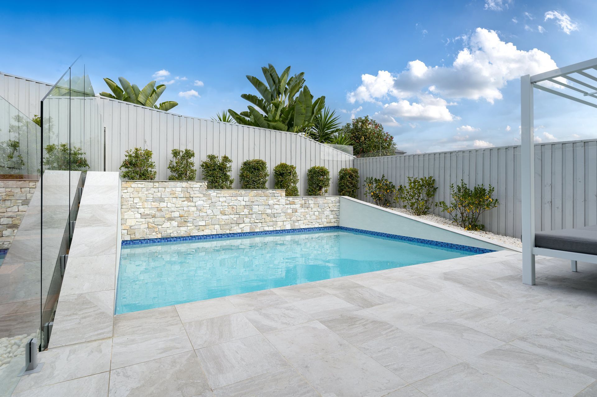 Small rectangular pool surrounded by light stone patio, white fence, blue sky.