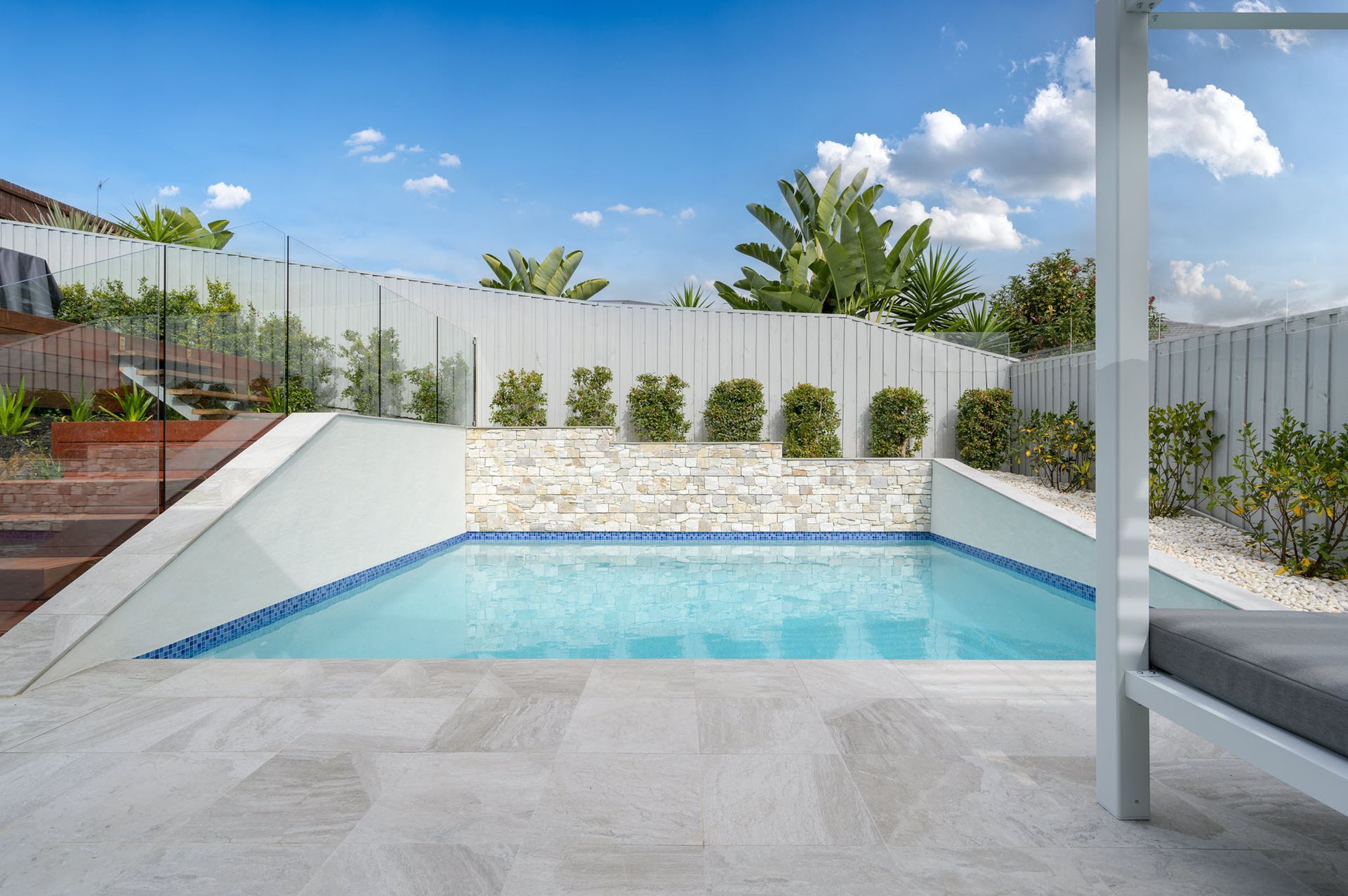 Small in-ground pool in a backyard with white walls, fence, and lounge area; blue water and sky.