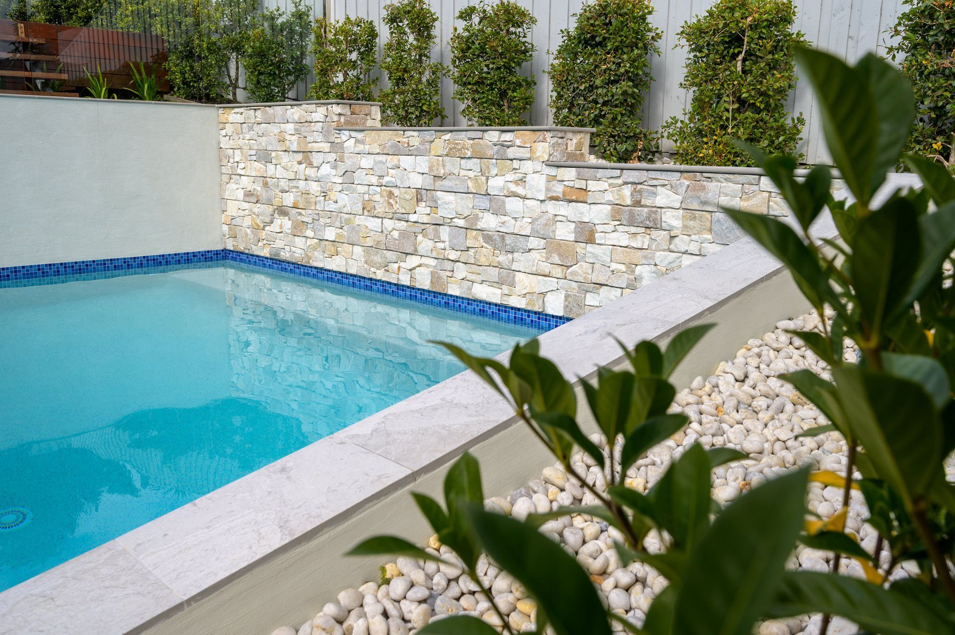 Pool with stone wall, greenery, and white pebbles.
