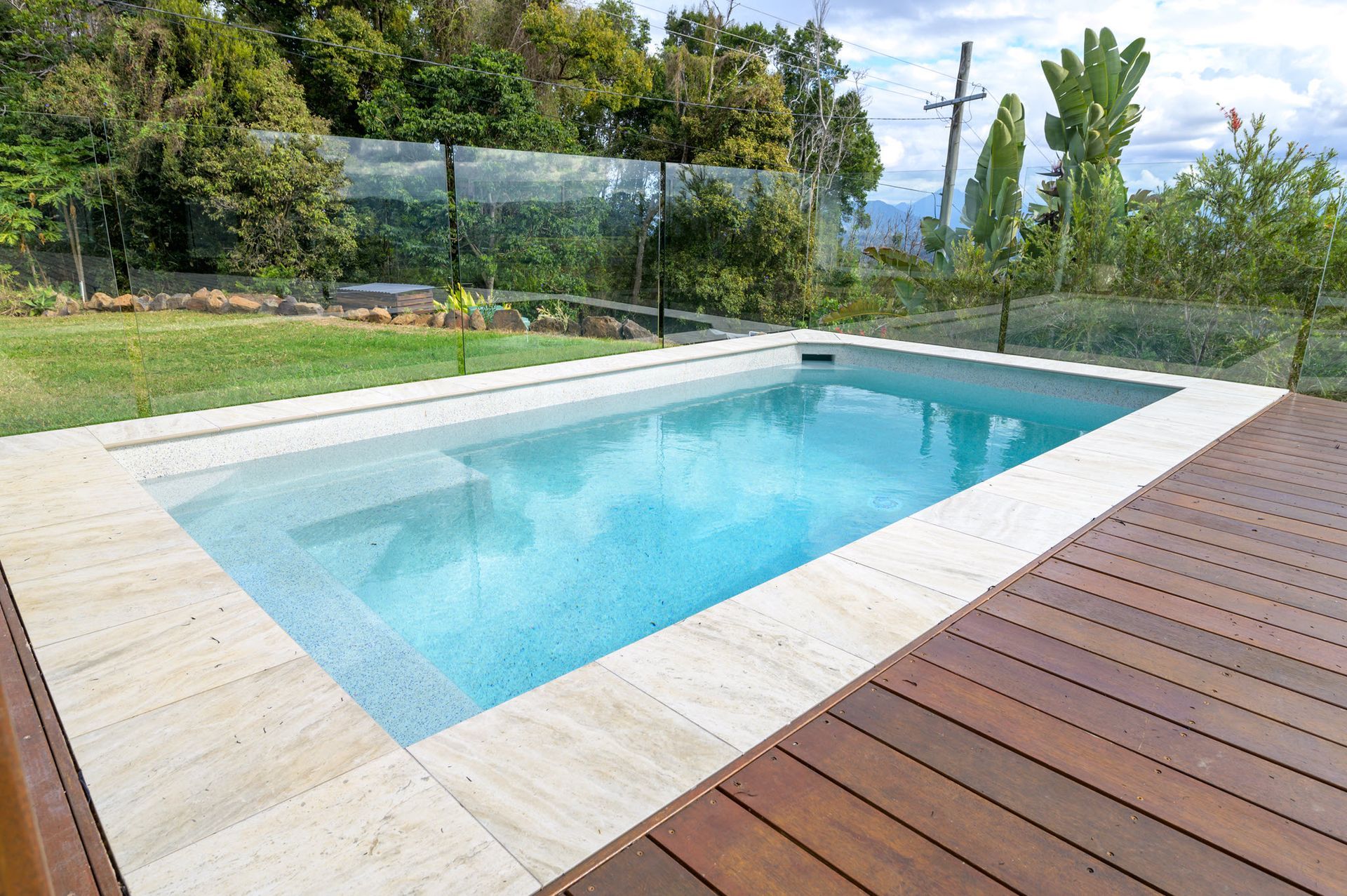 Rectangular pool with blue water and sandstone coping, surrounded by a wood deck and grass lawn.