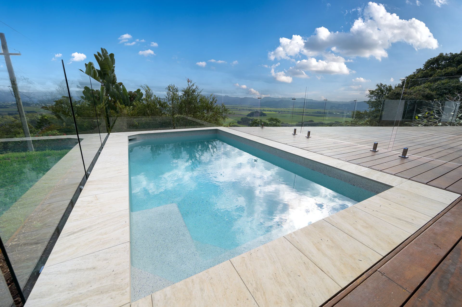 Rectangular pool with light-colored stone edging, surrounded by wood decking and glass railing, overlooking a scenic vista.