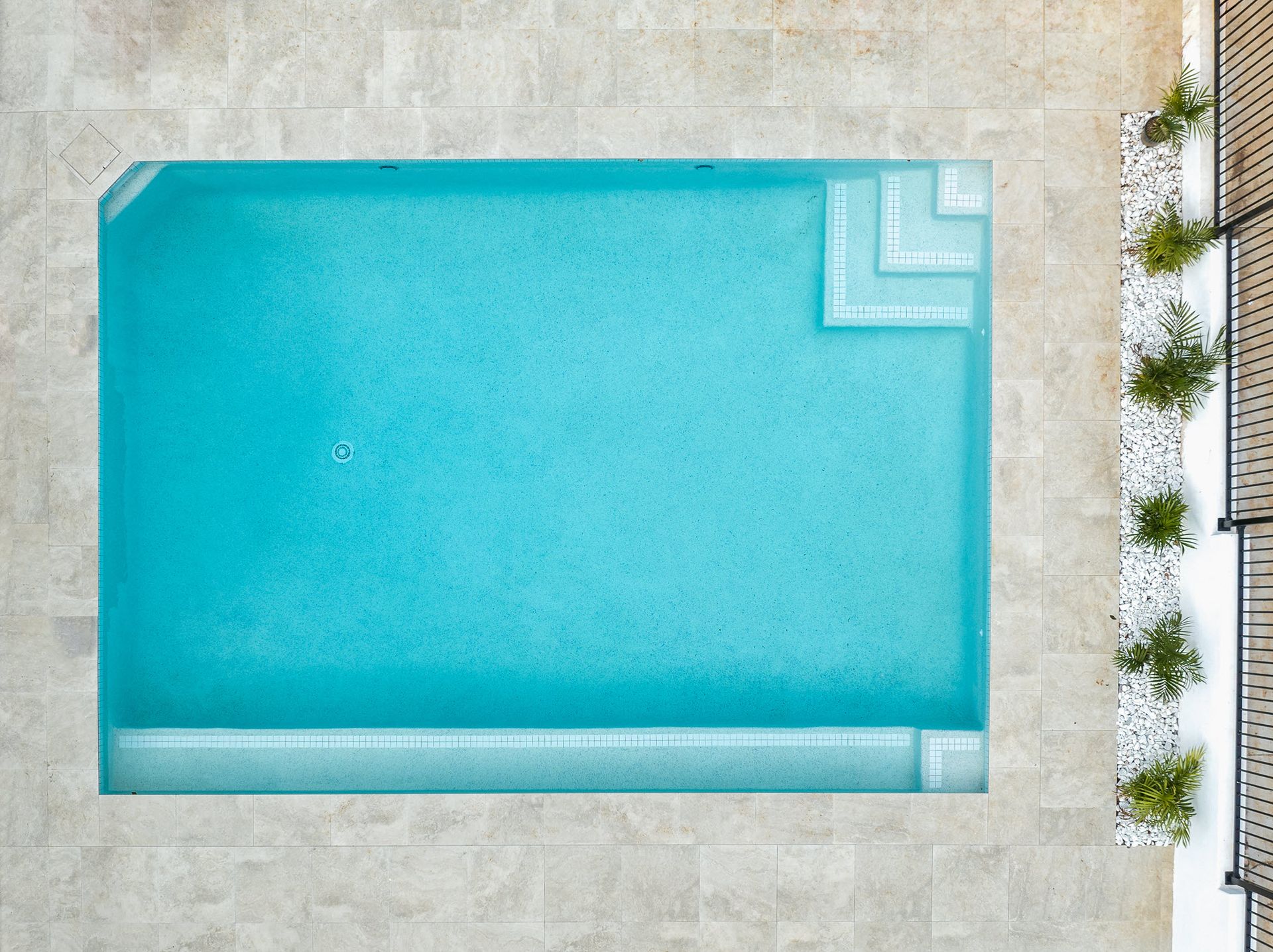 Overhead view of a turquoise pool with steps and a beige tiled surround, next to a white rock garden and black fence.