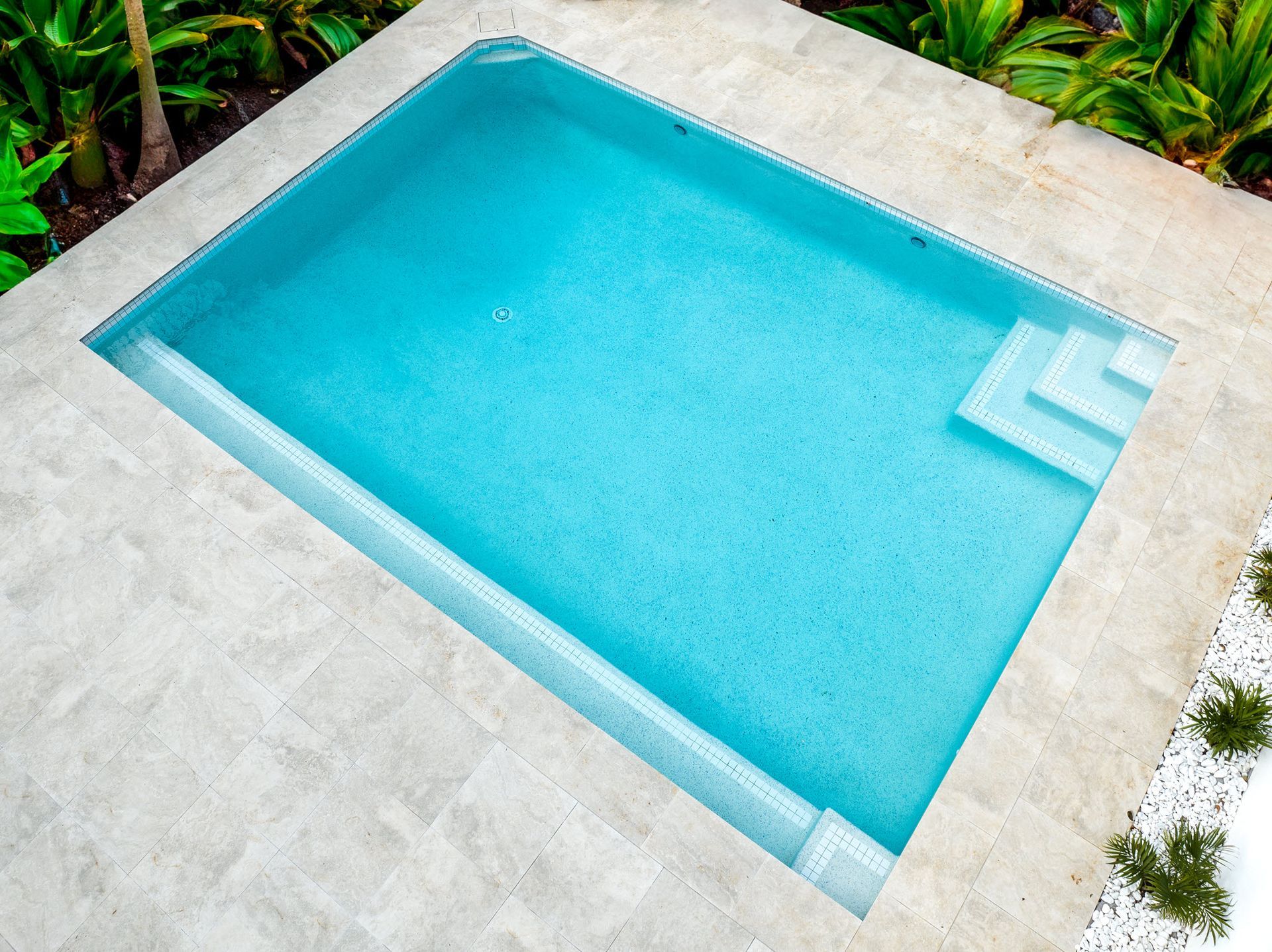 A small, rectangular swimming pool with light blue water, surrounded by light-colored stone tiles.
