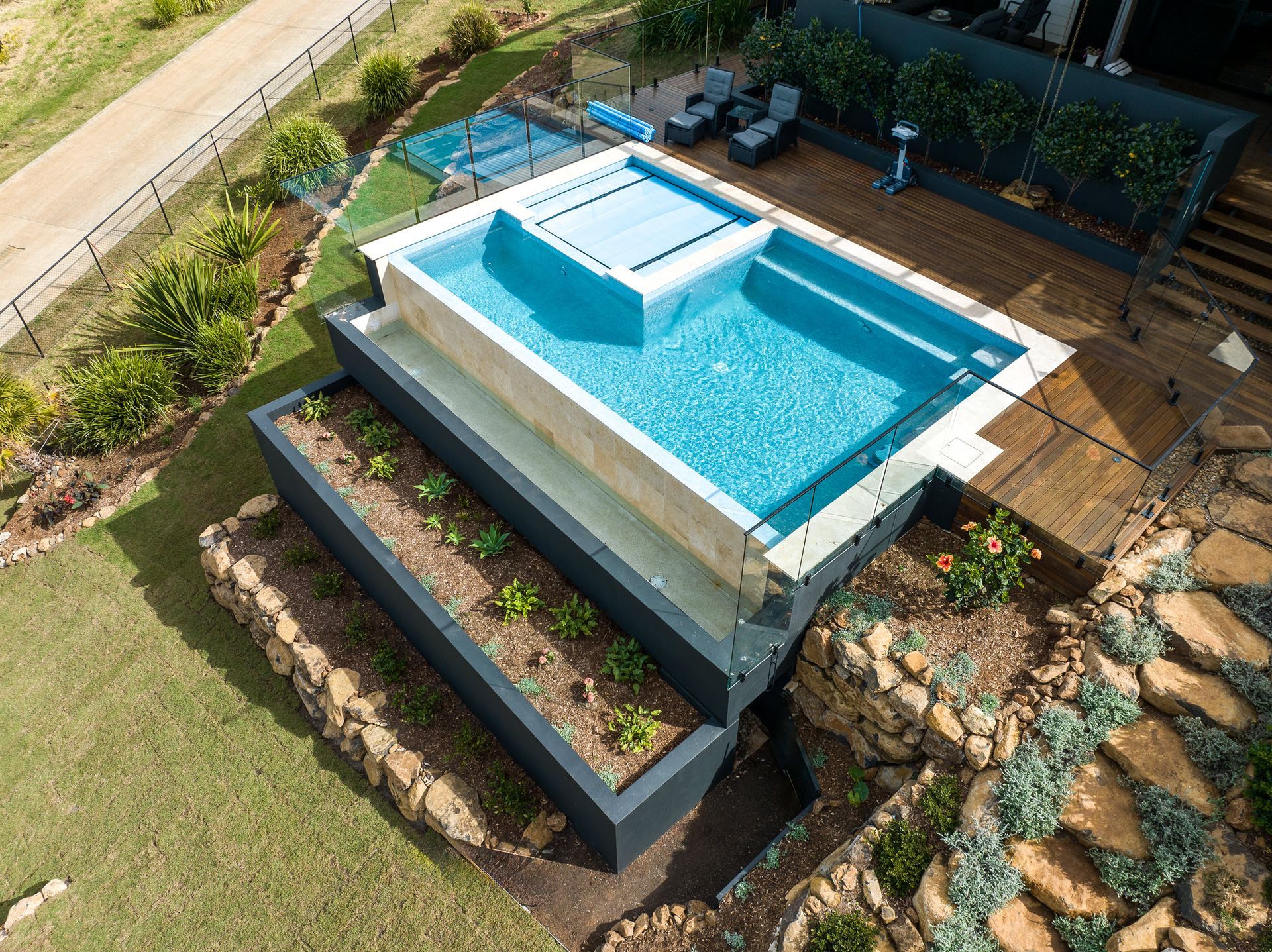 Aerial view: luxurious backyard pool with stone planters, wooden deck, and surrounding landscaping.