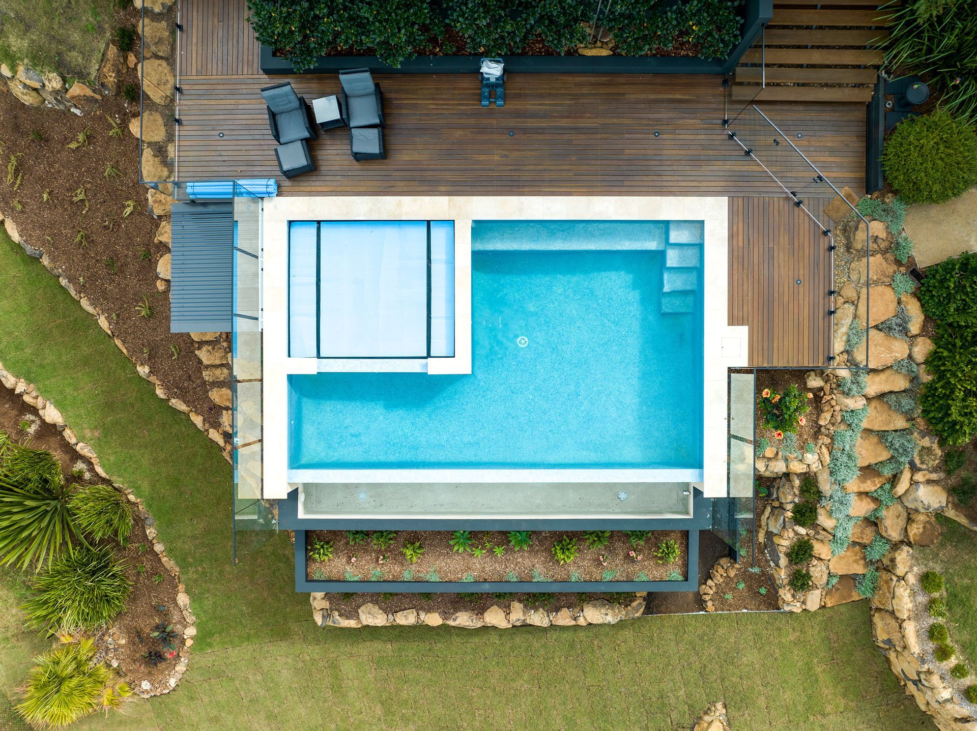 Aerial view of a rectangular swimming pool with a spa, surrounded by wooden deck, landscaping, and green lawn.