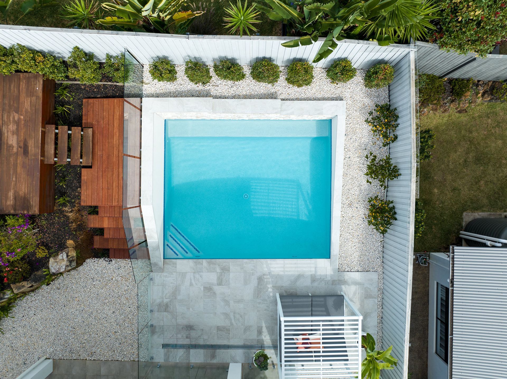 Overhead view of a square turquoise pool surrounded by a wooden deck, white fence, and gravel.