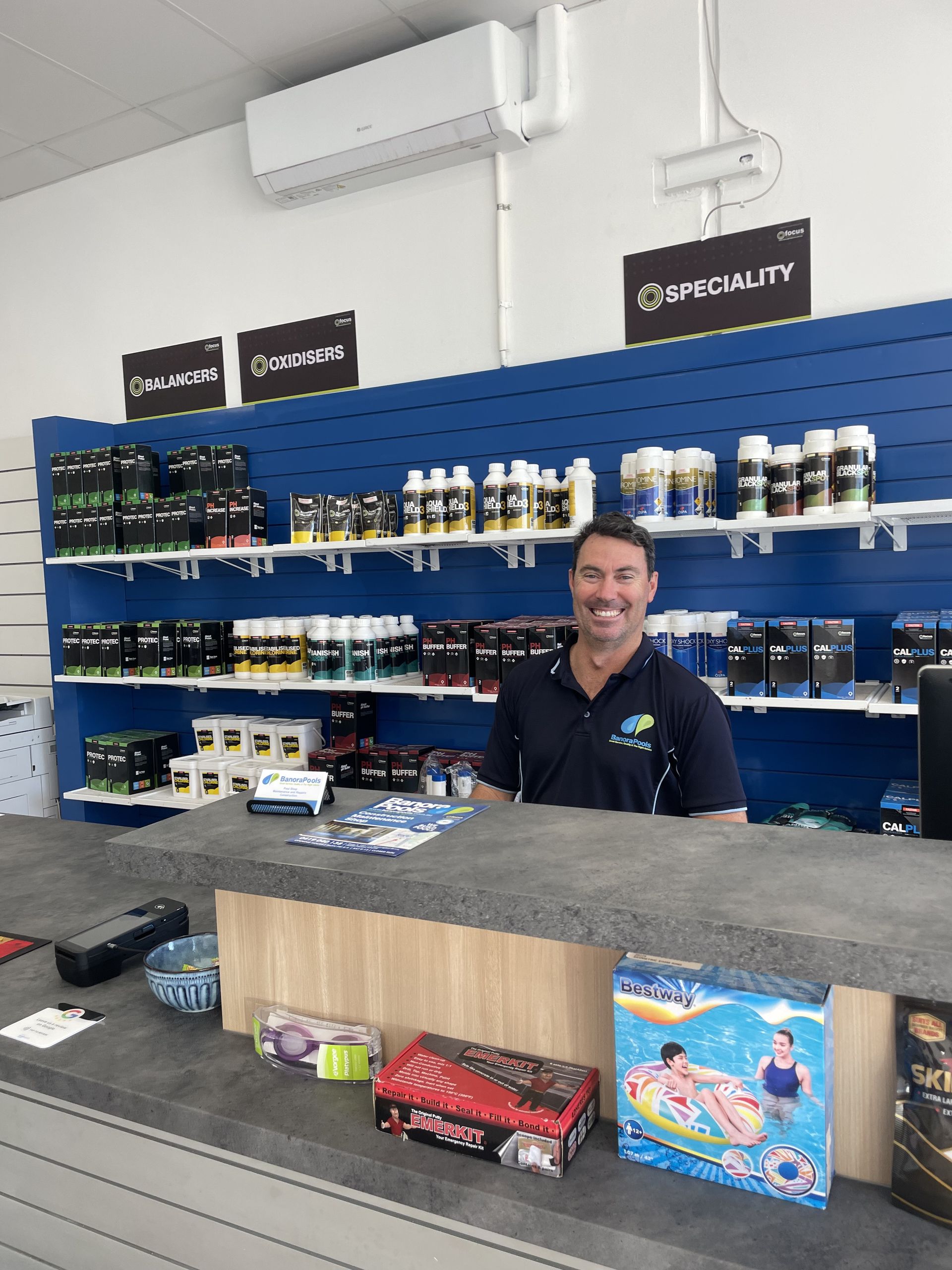 Man smiling behind a shop counter with products displayed on shelves, blue wall. — Banora Pools in Tweed Heads South, NSW