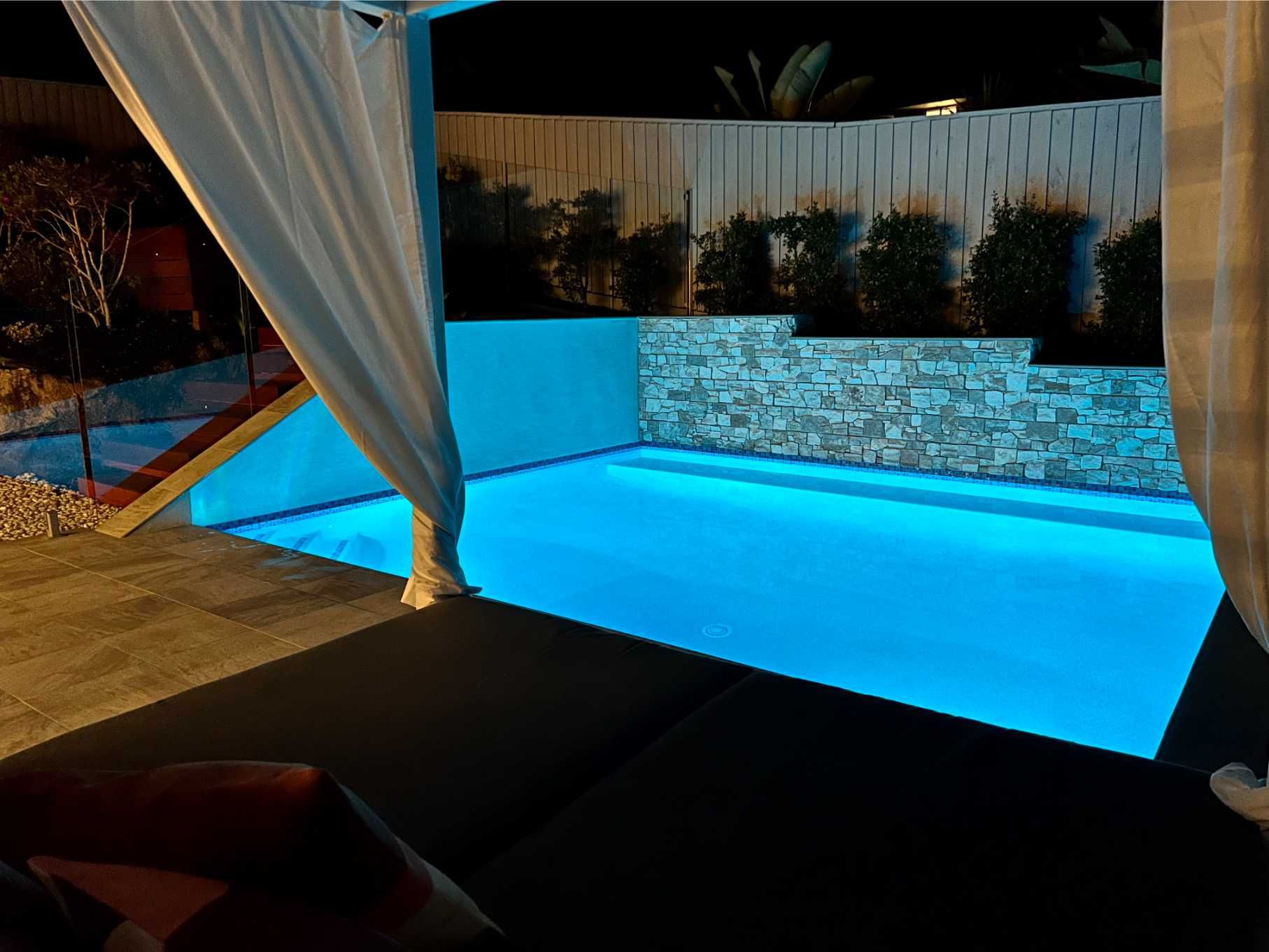 Night View of a Blue-lit Pool. White Cabana Curtains Frame the Scene. Stone Wall, Dark Patio, and Greenery — Banora Pools in Tweed Heads South, NSW
