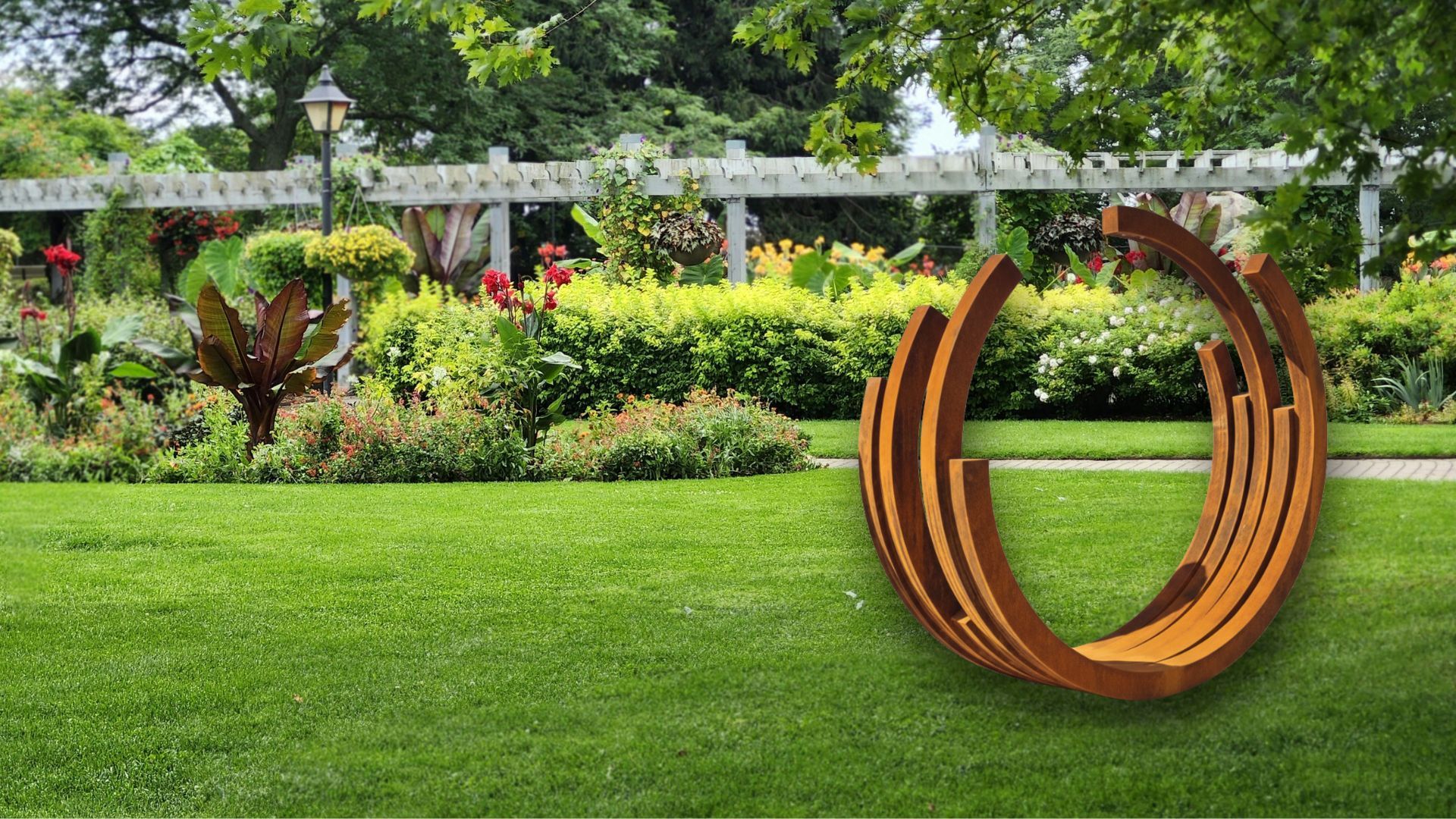 A circular, rust-colored metal sculpture sits on a green lawn in front of a garden with a trellis.