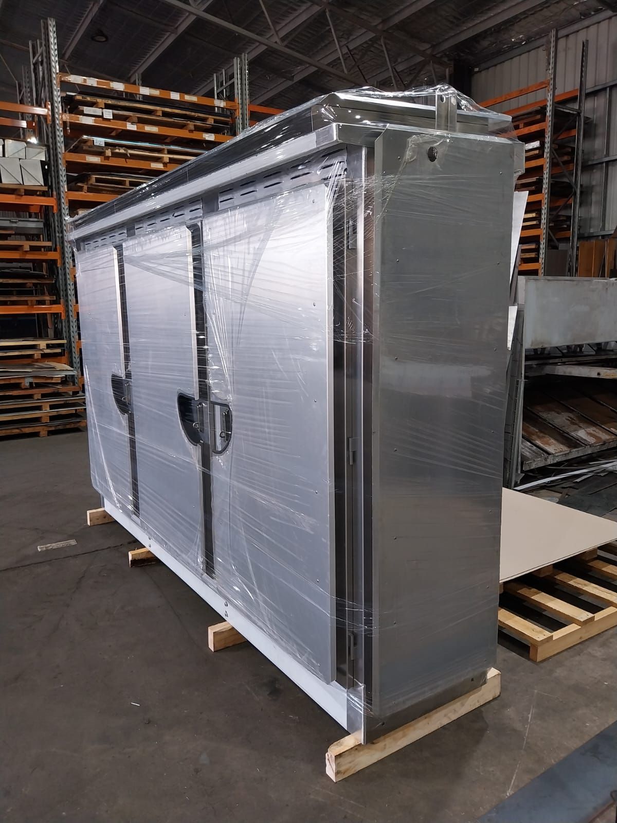 A new, stainless steel commercial refrigerator wrapped in plastic sits on wooden shipping pallets inside a warehouse.