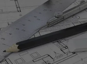 A black pencil, a metallic ruler, and a drafting compass rest on top of detailed architectural floor plan drawings.