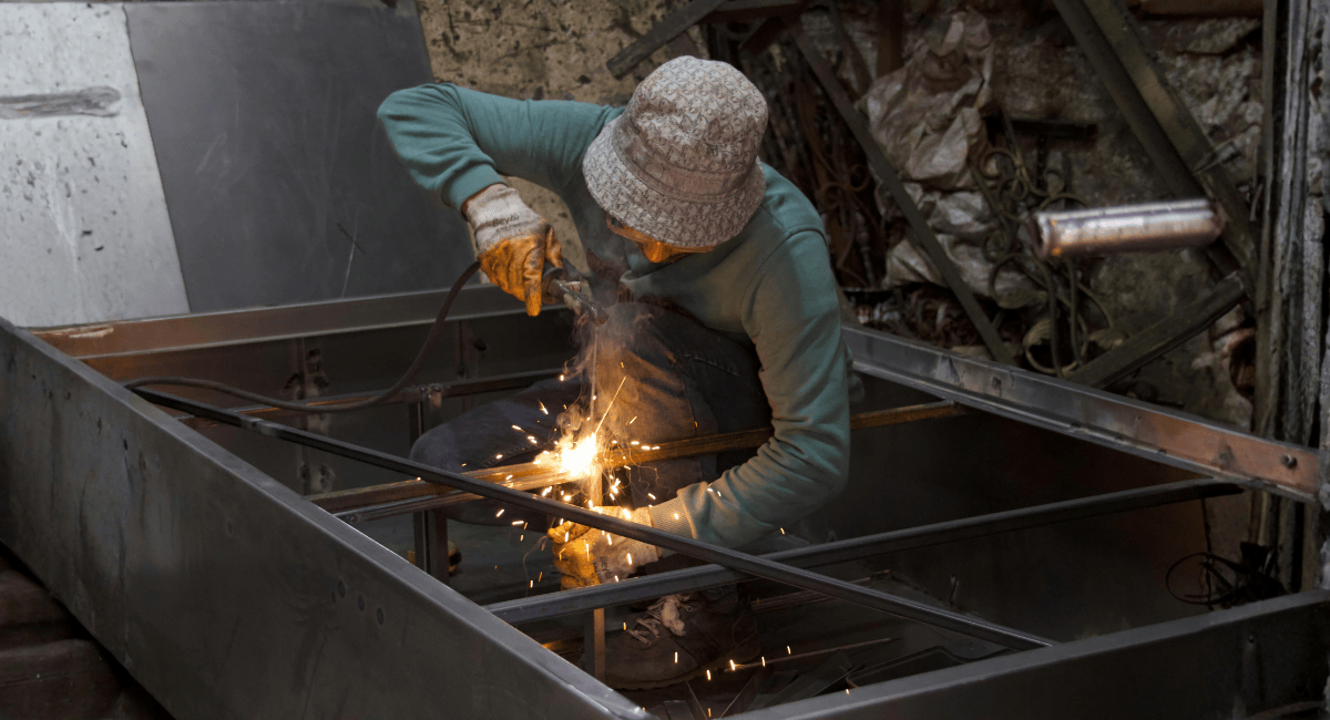 A worker in an orange hard hat and blue coveralls uses a laptop in a warehouse filled with stacks of metal pipes.