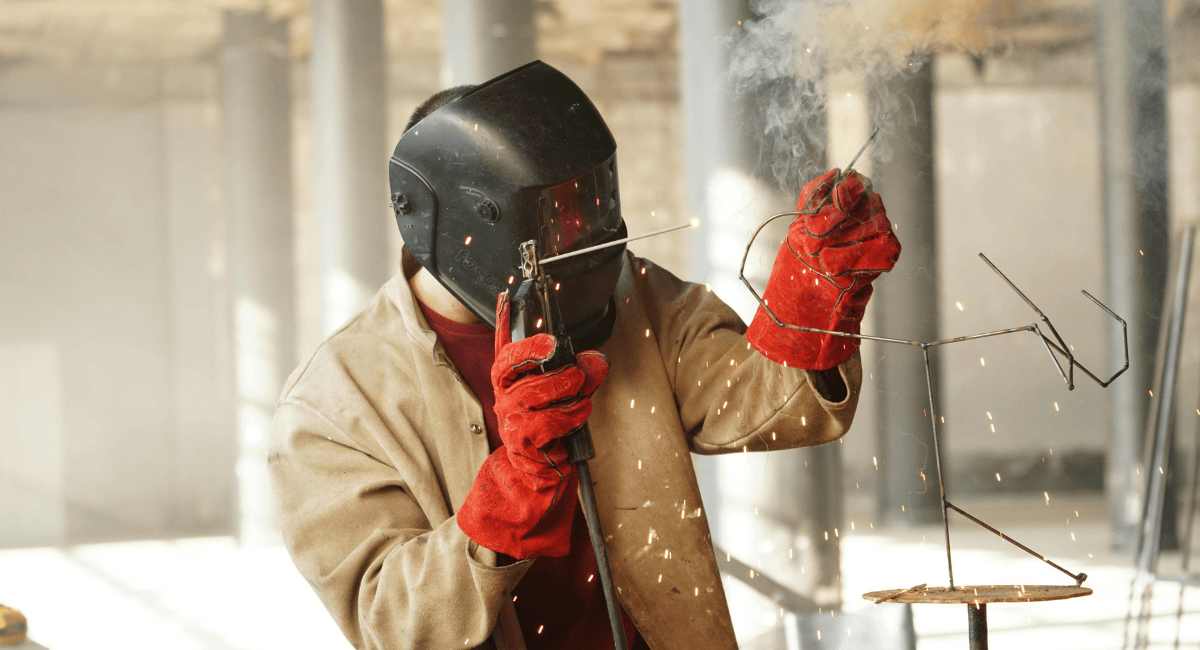 A welder wearing a mask and red protective gloves sparks a metal sculpture in a workshop.
