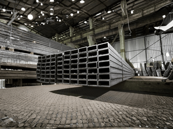 A stack of rectangular hollow steel beams resting on a patterned floor inside a large industrial warehouse.