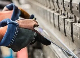 A gloved hand holds a thin sheet of metal into a mechanical press brake for bending in a factory setting.