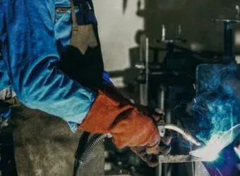 A welder in a blue uniform and orange protective gloves uses a torch to weld metal, creating sparks in a workshop.