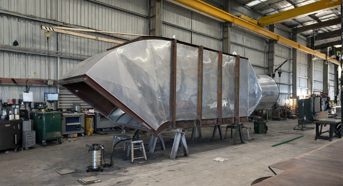 A large metal duct component with support ribs sits on stands inside a metal fabrication workshop.
