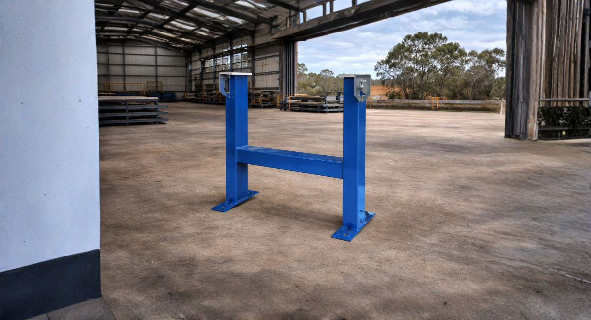 A blue, H-shaped steel frame stand sits on a concrete floor inside a large, open-walled industrial warehouse.