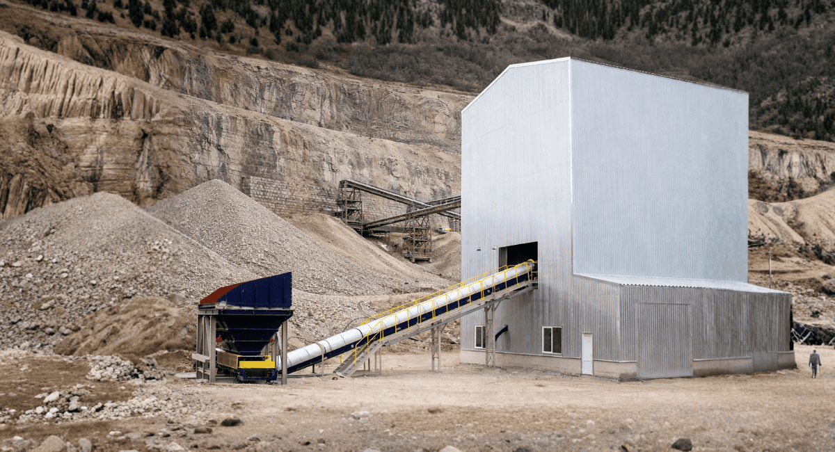 A conveyor belt feeds rock from a blue hopper into a large, silver industrial building in a stone quarry.