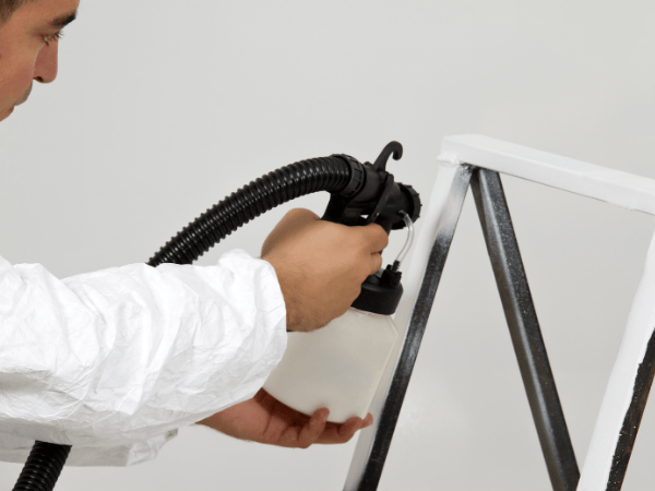 A person wearing protective clothing uses a paint sprayer to coat a metal frame white.
