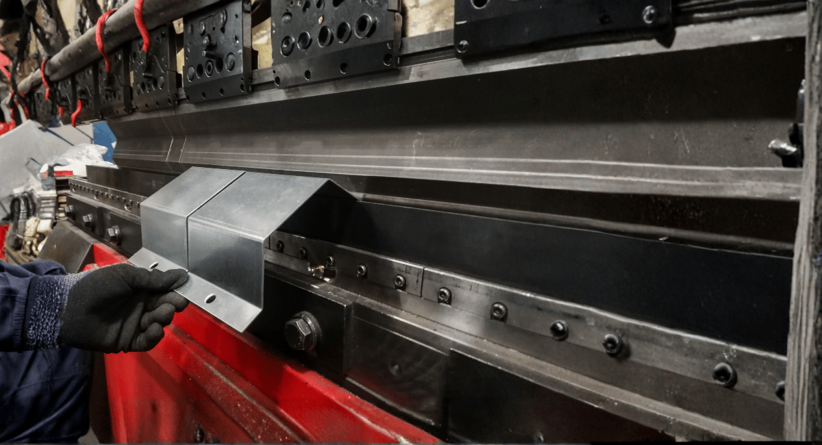 Metal worker bending metal on a large industrial press. The bent metal has a roof-like shape.