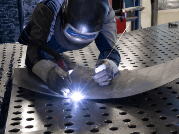 Welder in a workshop, welding metal sheet on a perforated table; sparks fly.