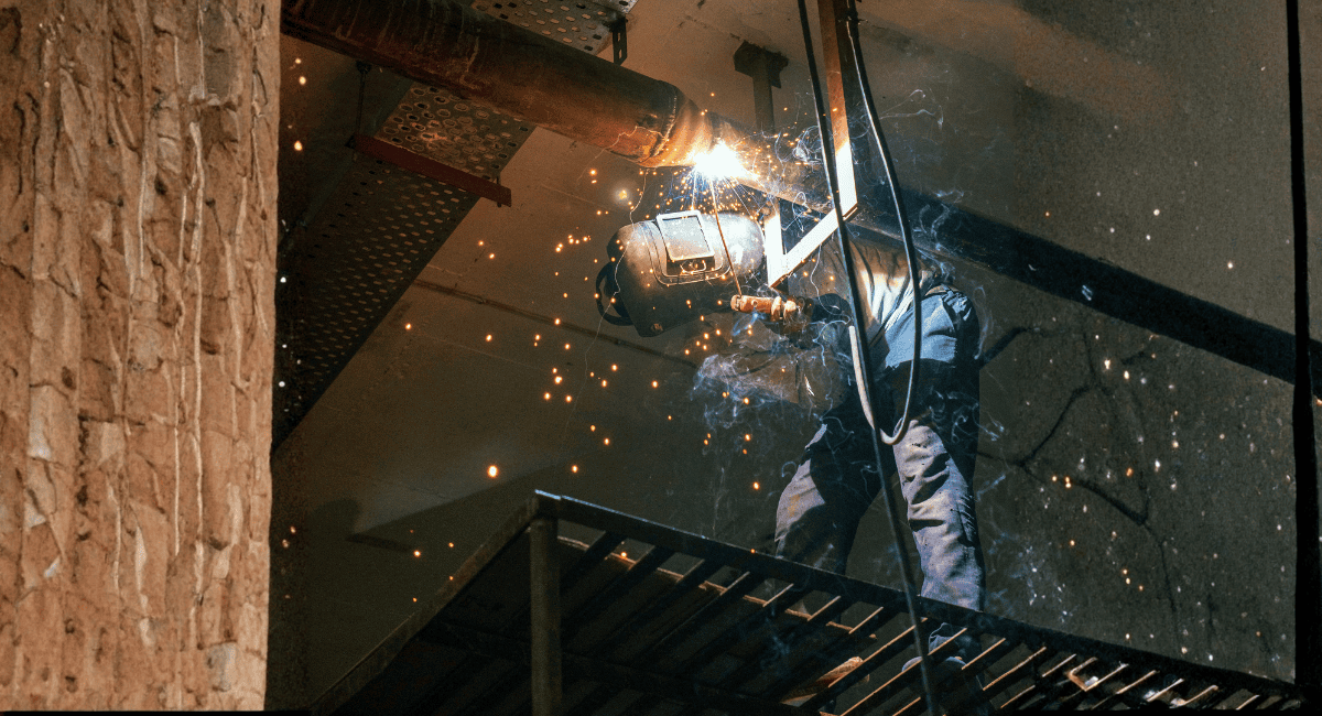 A person wearing protective gear performs overhead welding on a metal pipe, creating bright sparks in an industrial setting.