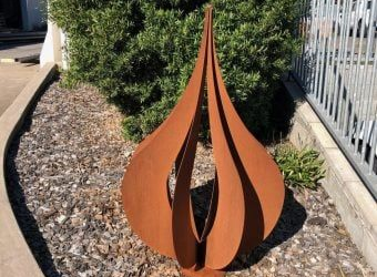 A rust-colored, teardrop-shaped metal sculpture stands in a gravel garden bed in front of a green bush and metal fence.