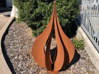 A brown, rusted metal sculpture with a flame-like, teardrop shape placed in a garden bed with gravel and green foliage.