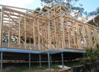 A wooden house frame under construction, supported by metal posts on a sloped site with trees in the background.