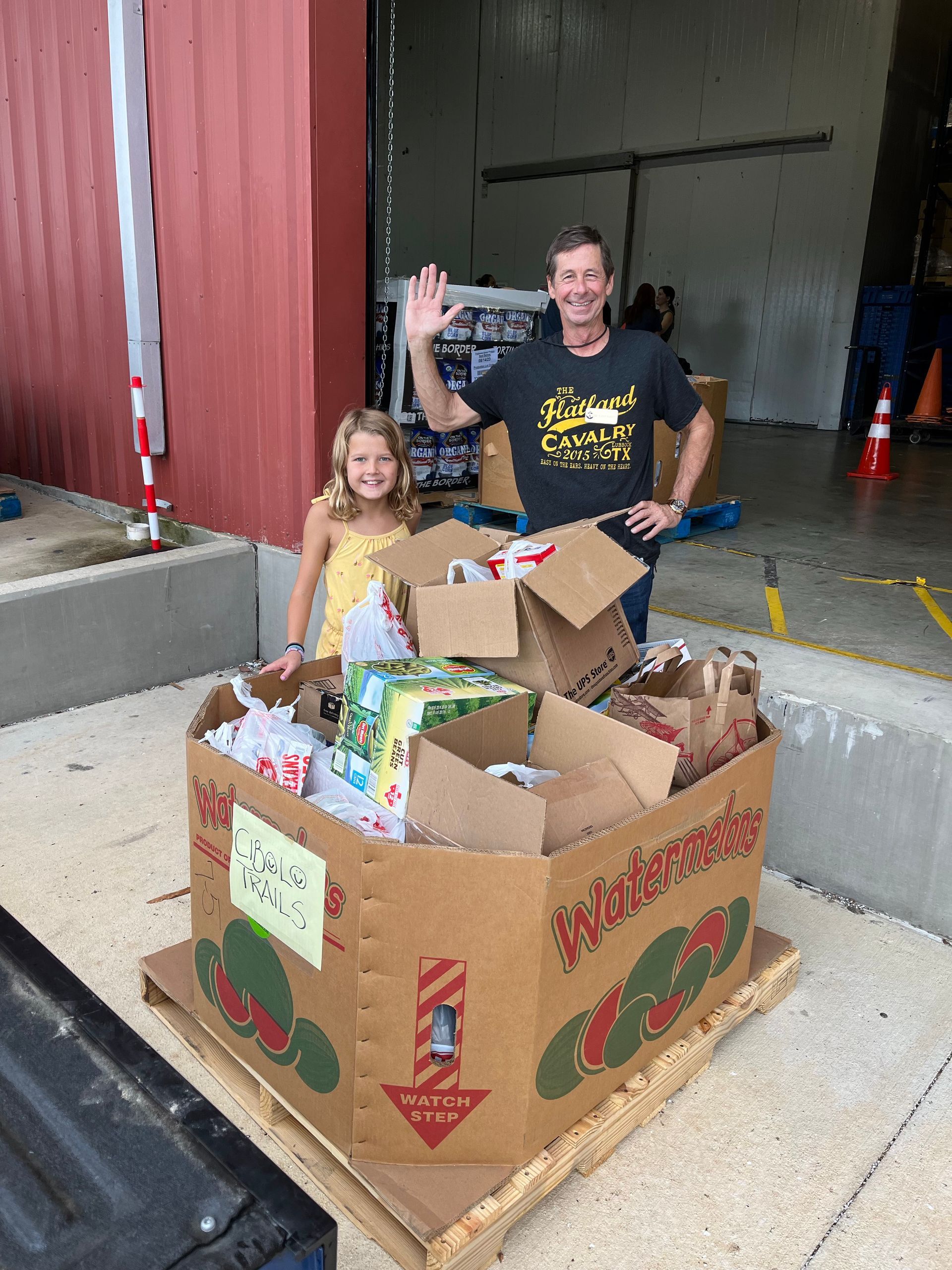 A man and a little girl are standing next to a cardboard box filled with food.