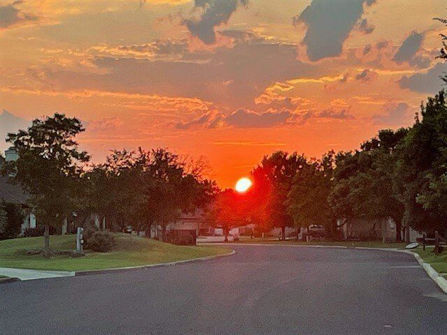 The sun is setting over a residential area with trees in the foreground.