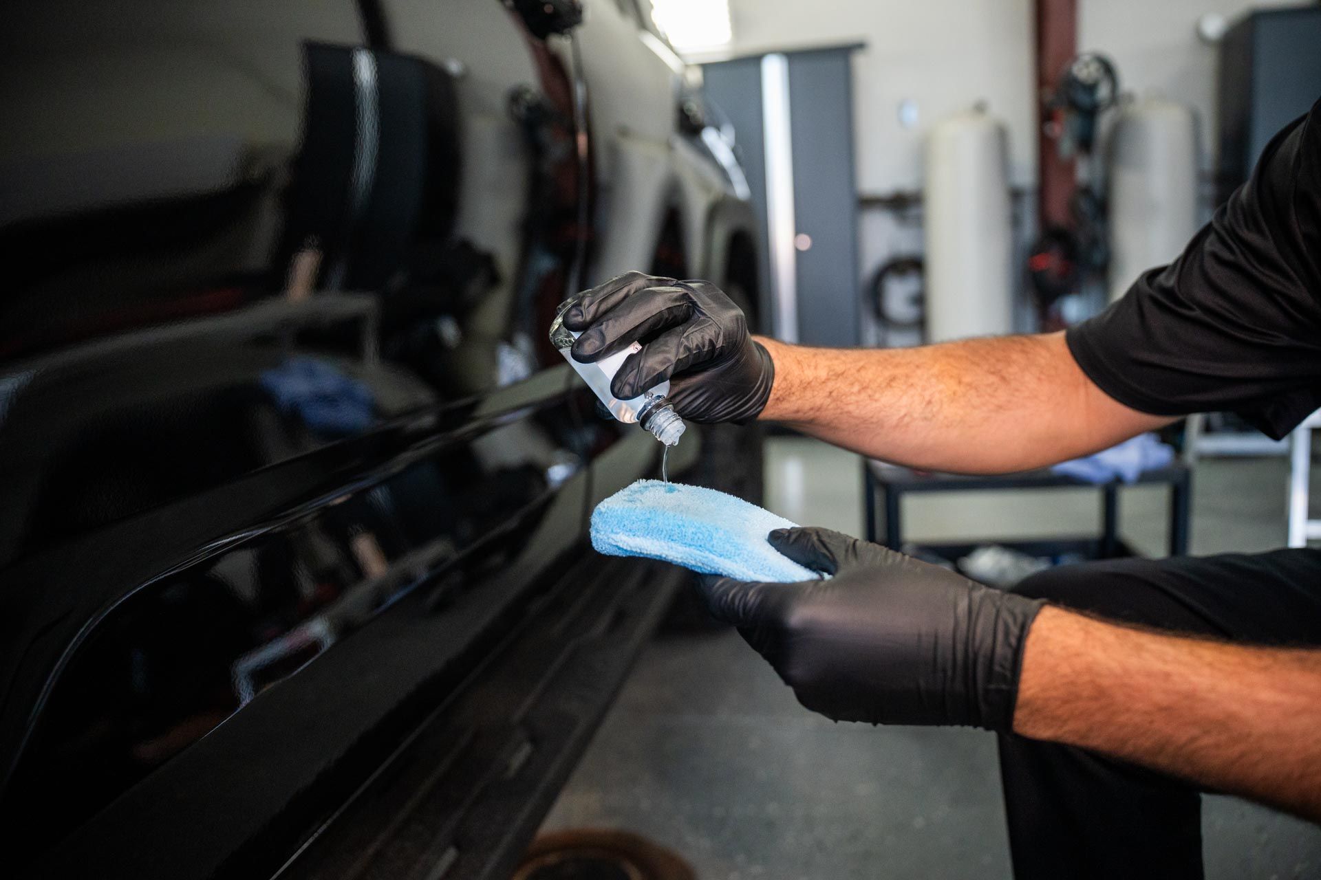 A man wearing black gloves is cleaning a black car with a blue cloth.