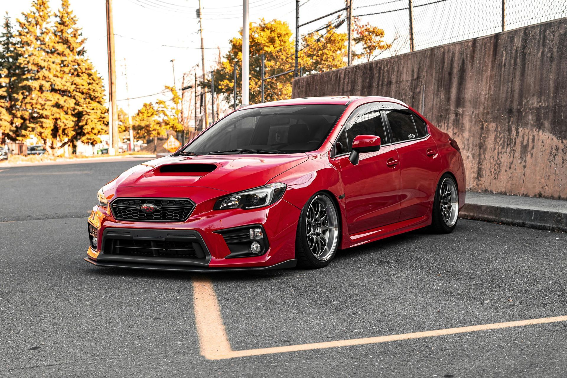 Red Subaru WRX sedan parked on asphalt next to a concrete wall; trees and a fence are in the background.