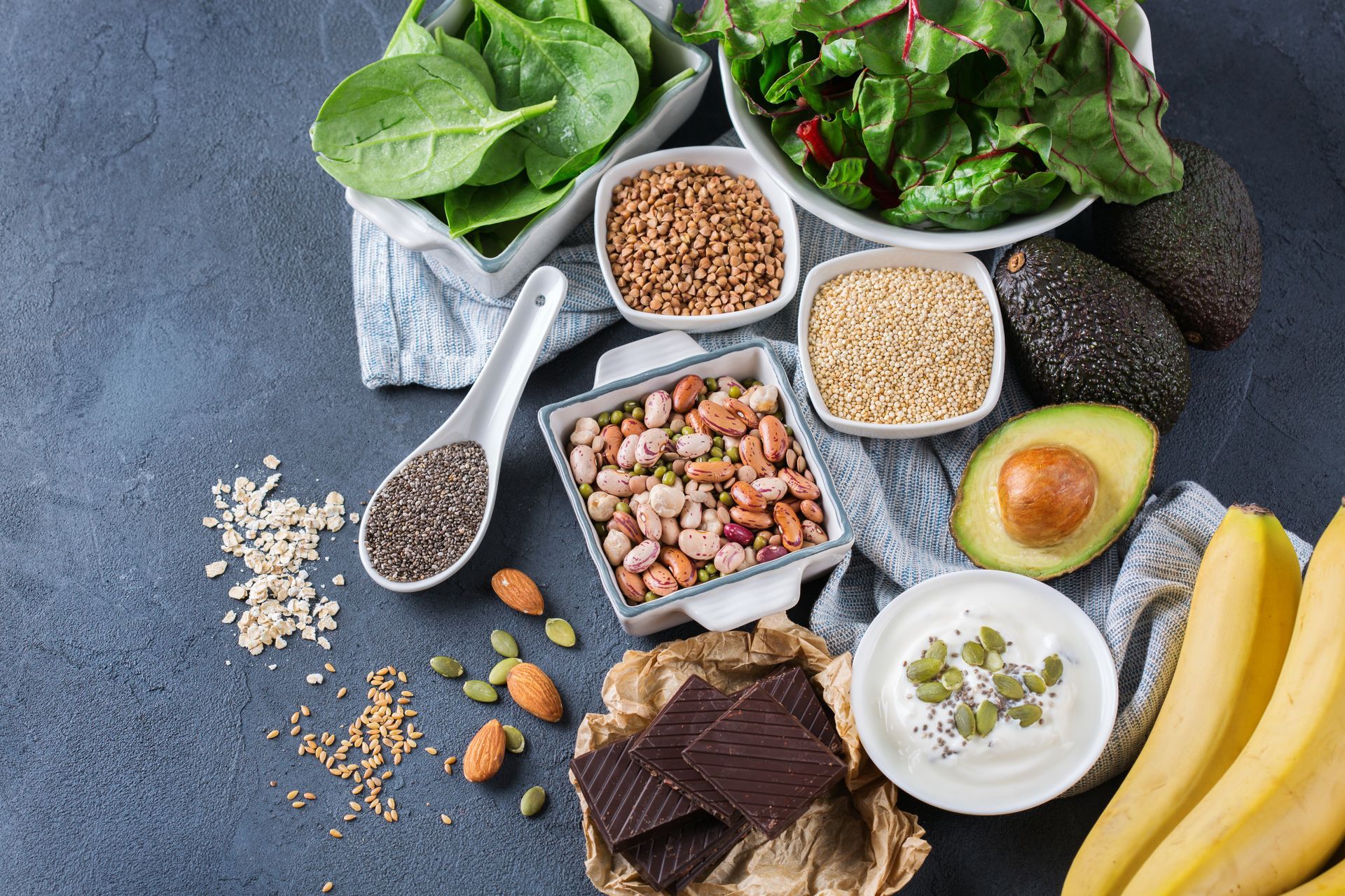Assortment of healthy foods: spinach, salad, nuts, seeds, avocado, yogurt, bananas, and dark chocolate.