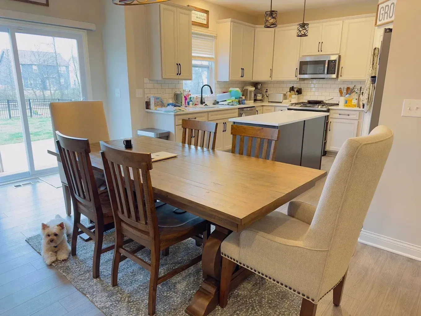 A dining room with a wooden table, chairs, and a small dog on a rug. Kitchen in the background.