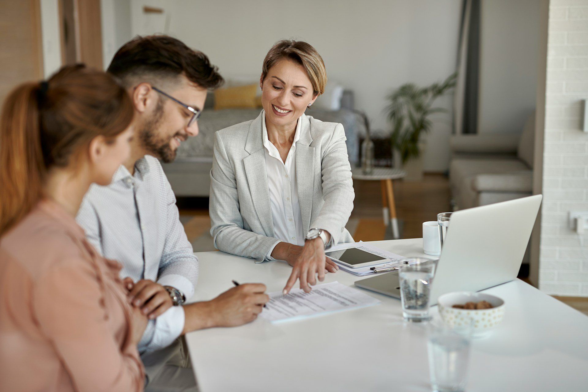 A professional advisor gestures toward a document for a couple to sign at a table with a laptop.